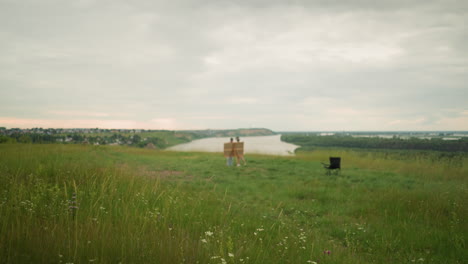 in a green field of tall grass with wildflowers, the background is intentionally blurred, revealing a serene lakeside scene. two distant figures and a lone chair near the lake