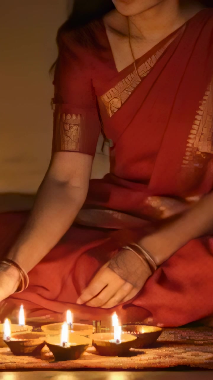 Vertical video: Using lit wick, woman in red sari lighting diyas on patterned mat, offering prayer