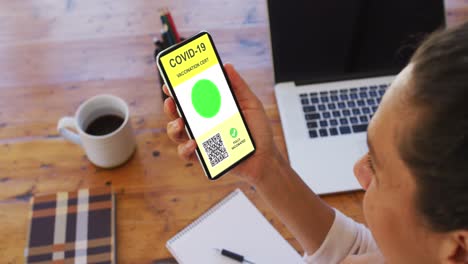 Woman-at-desk-holding-smartphone-with-covid-vaccination-certificate-and-qr-code-on-screen