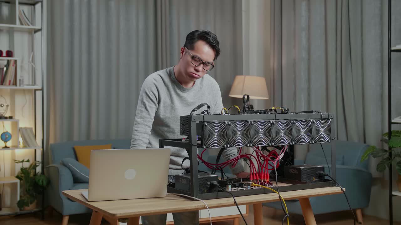 안경을 쓴 아시아인 남자가 머리를 흔들고 노트북과 광산 기계를 사용하여 암호화폐를 채굴하는 동안 두통을 고 있습니다.