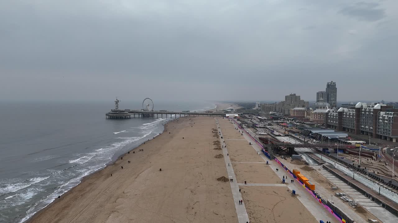 Walking tourist on sandy beach and Promenade of Scheveningen Town during cloudy day. Aerial forward wide shot. Ferris Wheel and pier in distance. Netherlands, Europe at grey cold winter day.