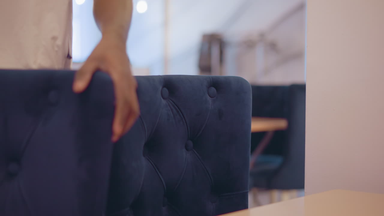 Close-up shot of tufted blue chair with person in white shirt standing in background, focused on chair's detailed upholstery in modern indoor setting, highlighting design features