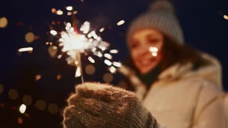 montage of a smiling woman wearing winter clothes, a beanie and mittens, holding a burning sparkler at night, with blurred lights in the background