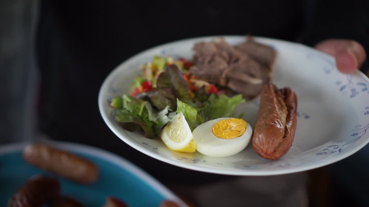 cámara lenta - el hombre pone salchichas y hamburguesas en su plato saludable con huevos y ensalada