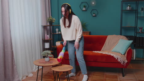 Young-dancing-woman-dusting-furniture-alone-caring-for-hygiene-using-colorful-duster-in-room-at-home