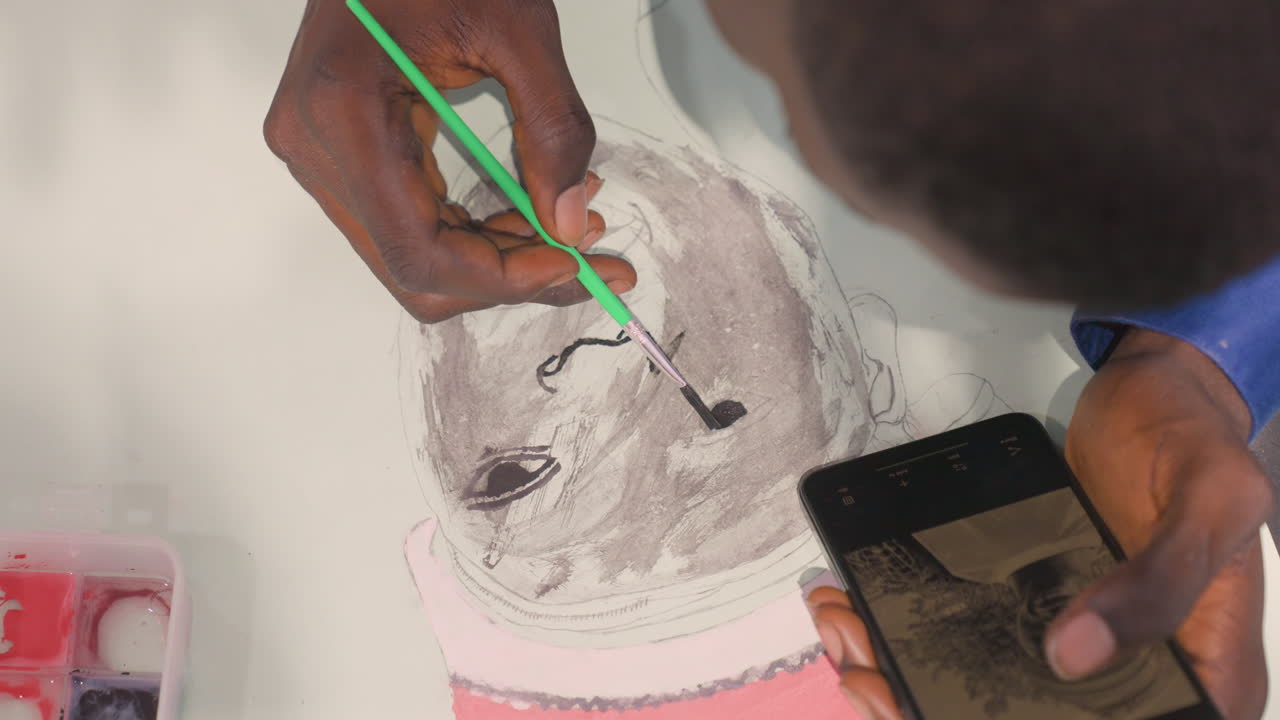 Black artist painting fine portrait details, overhead closeup of brush and phone reference, precise strokes on grayscale face, careful fingertip control and quiet focused atmosphere