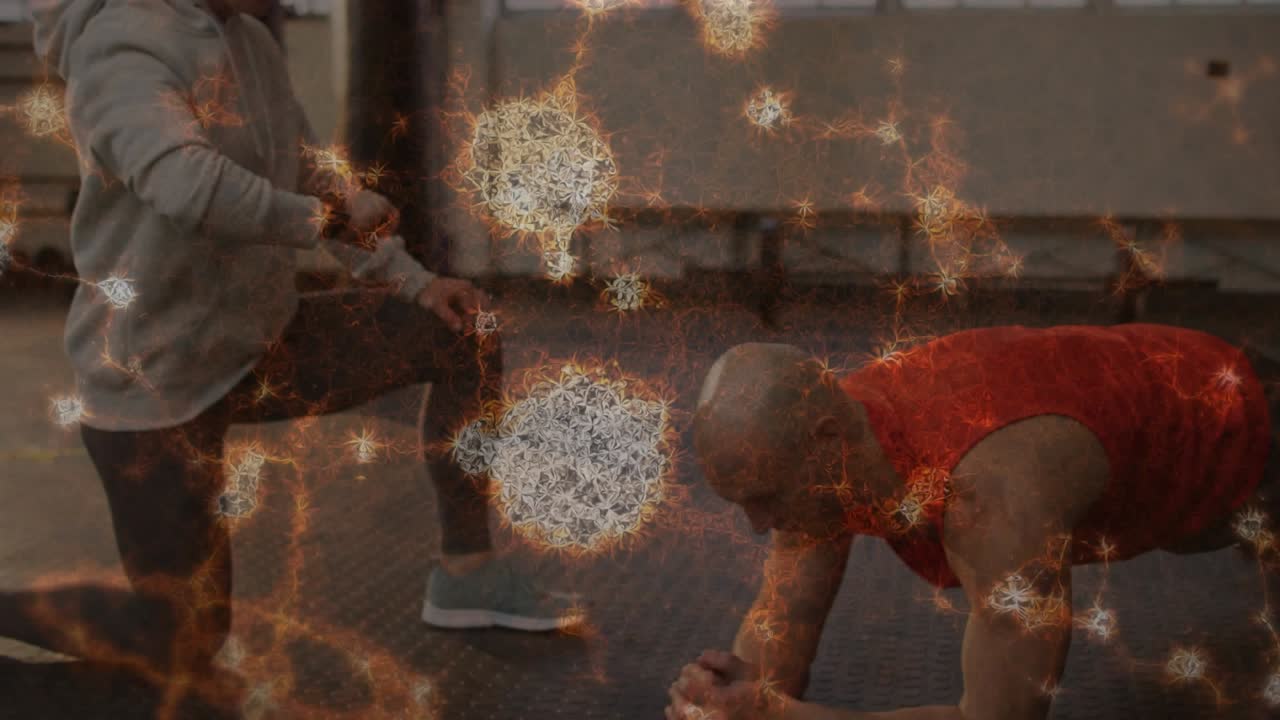 Coach kneeling starting timer while man holding plank in gym, particle overlay drifting over bodies