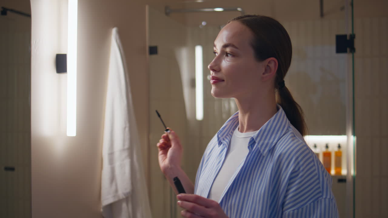 Ragazza rilassata che applica il mascara allo specchio del bagno. Donna sorridente che ammira la sua bellezza