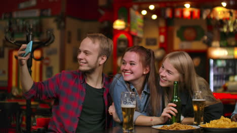 Un-Grupo-De-Amigos,-Un-Hombre-Y-Dos-Chicas,-Se-Toman-Selfies-En-Un-Bar-Riendo-Y-Sonriendo.-Pasar-Tiempo-Con-Tontos-En-Un-Bar