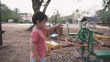 two year old asian boy enjoying playing with playground equipment pushing a merry go round at an outdoor park