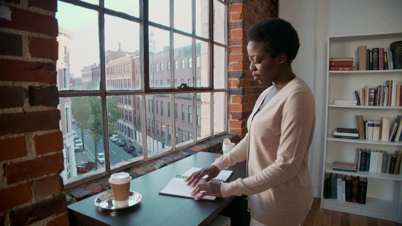 Opening spiral notebook, woman in beige top at window desk flipping pages checking notes