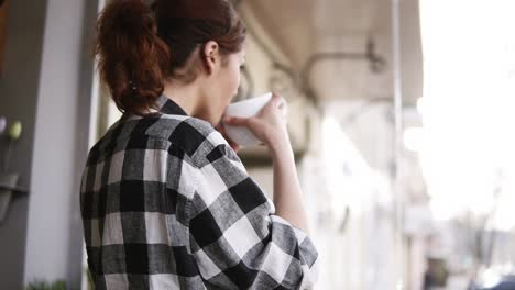 Una-Chica-Encantadora-Y-Complacida-Con-Una-Camisa-A-Cuadros-Se-Encuentra-Frente-A-La-Ventana,-Bebe-Un-Refresco-En-Una-Gran-Taza-Blanca-Y-Mira-Por-La-Ventana.-Atrás.-Y-Metraje-Lateral.-Perspectiva-Borrosa