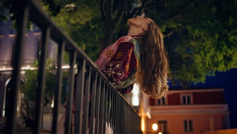 modelo de moda inclinado a las barandillas posando en el parque de la ciudad del anochecer. mujer de pie en la ciudad de la noche.
