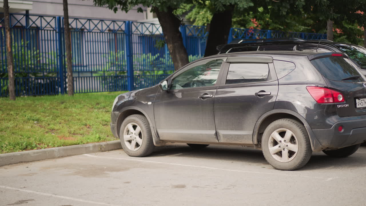 SUV grigio parcheggiato vicino a una recinzione, uomo apre la porta posteriore e scende sul marciapiede illuminato dal sole, ruote infangate, ombra di albero frondoso, scena urbana a bordo strada, jeans casual, giacca rossa, arrivo di un pendolare quotidiano ripreso candidamente.