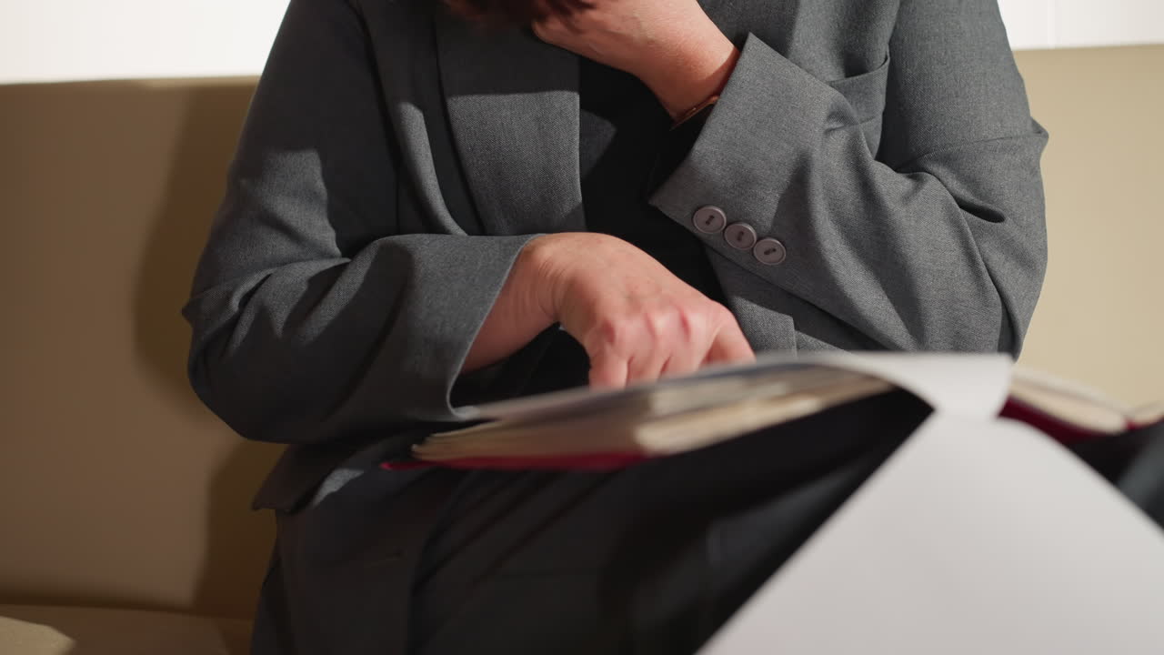 Businesswoman in gray suit reading document while sitting, pointing finger to page with thoughtful expression, possibly preparing for presentation or meeting, showing focus and concentration