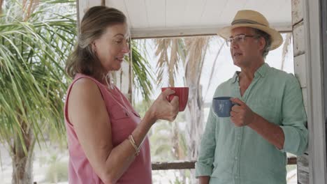 Happy-senior-caucasian-couple-drinking-coffee-and-talking-on-porch-of-beach-house,-in-slow-motion