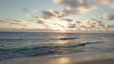 Aerial-shot-of-an-ocean-at-sunrise-revealing-a-sandy-beach-and-a-lifeguard-house-with-clouds-in-the-sky