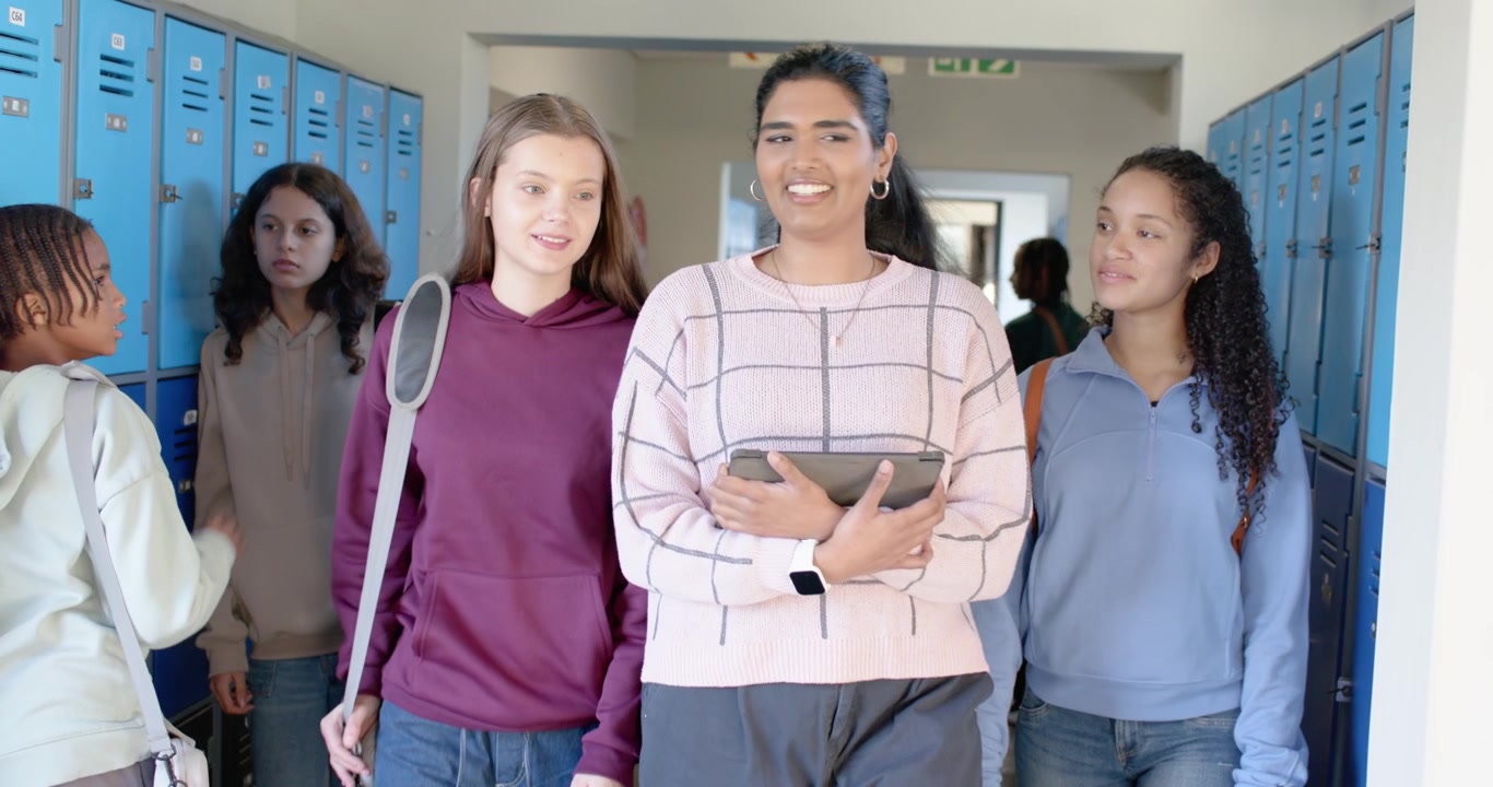 Diverse compagne di classe adolescenti camminano tra le lezioni a scuola, passando davanti agli armadietti blu e tenendo un tablet