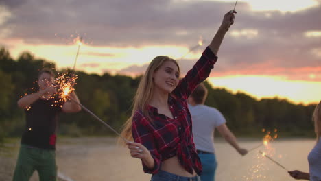 the girl in red plaid shirt and denim shorts is dancing with big bengal lights in her hands on the sand beach with her friends. this is beautiful summer evening on the open air party.