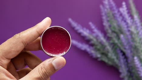 hand holding a small tin of red lip balm with lavender background