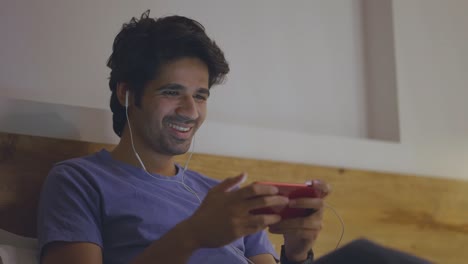 young happy indian asian male sitting on bed looking into mobile phone at home
