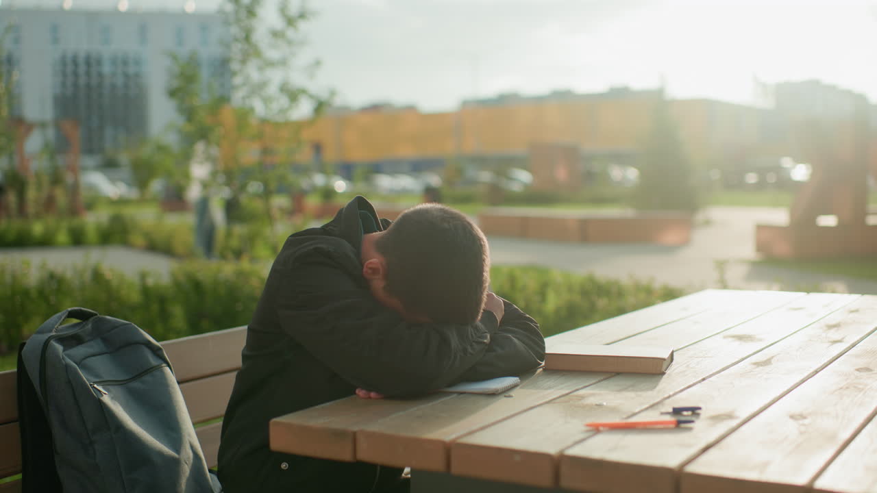 Boy sits at outdoor wooden table writing in notebook, accidentally drops pen, then leans forward resting head on table with thoughtful tired expression in calm daylight park atmosphere