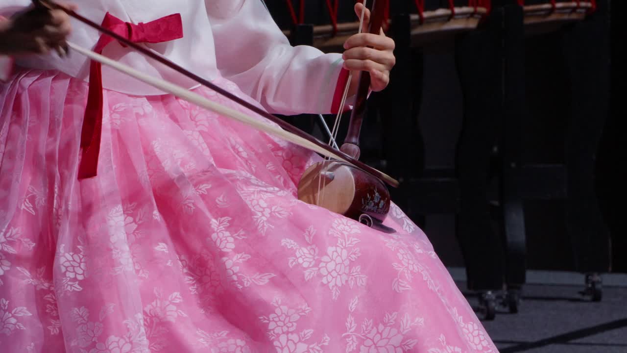 A Korean woman in a pink hanbok plays the haegeum, a two-stringed fiddle, during a traditional music performance. Close-up shot