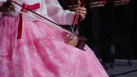 a korean woman in a pink hanbok plays the haegeum, a two-stringed fiddle, during a traditional music performance. close-up shot