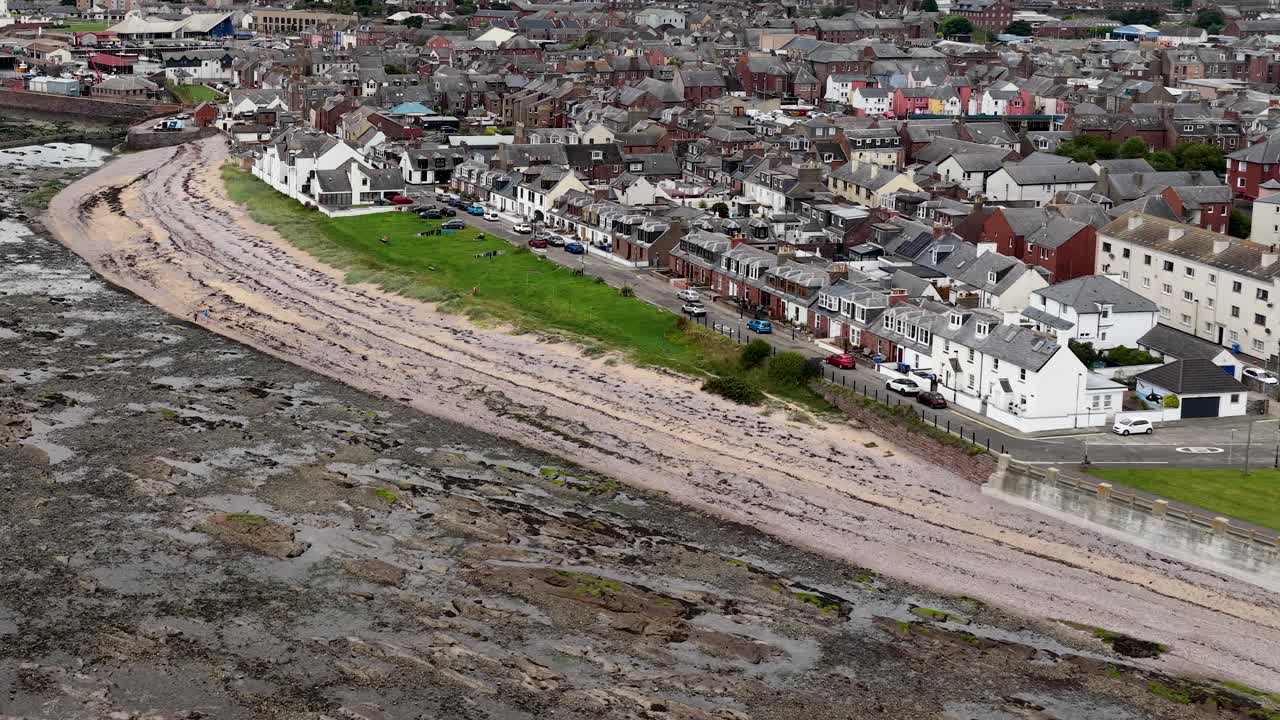 Drone pans above Scottish coastal town, revealing rocky shoreline, beachfront, and overcast skies