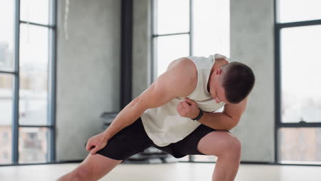 man performing side lunge warm up exercise in bright modern gym wearing white tank and black shorts balancing on light wooden floor under hanging straps by large windows in urban studio, controlled