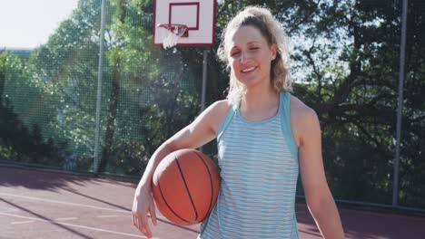 Portrait-of-caucasian-female-basketball-player-holding-ball-on-sunny-court,-in-slow-motion