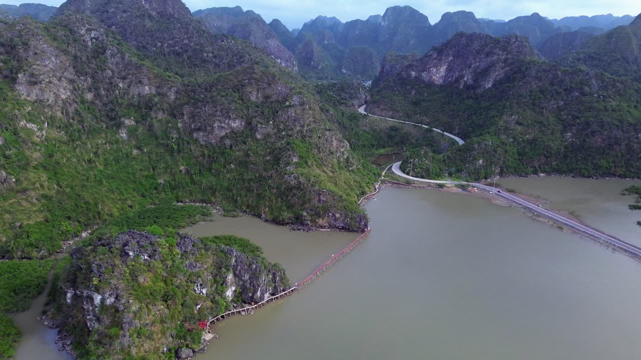 Drone zooms in over a winding mountain road near a muddy river on a cloudy day in Cat Ba Island