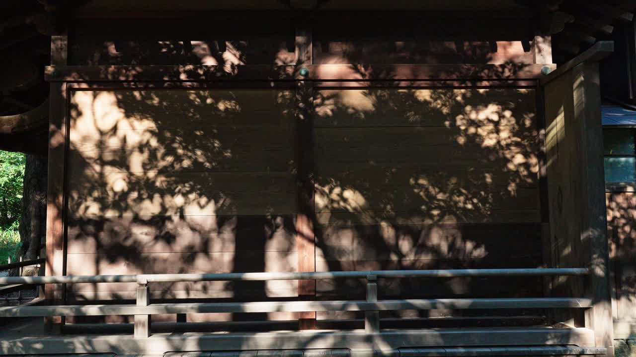 The weathered wooden facade of a traditional Japanese shrine is dappled with shadows from nearby trees under a bright sky.