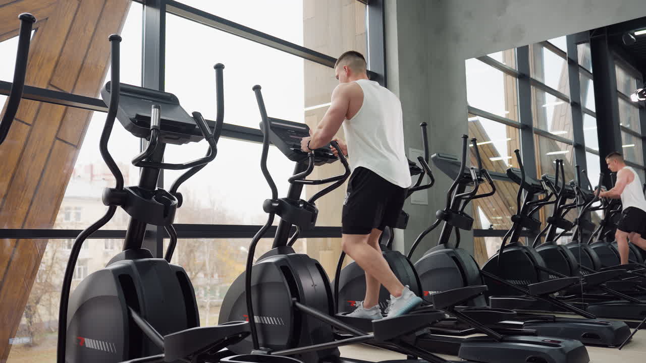 Young handsome man works out on elliptical trainer in sleek urban gym, adjusting settings on console, mirror reflecting him and others during energetic cardio training session beneath tall windows