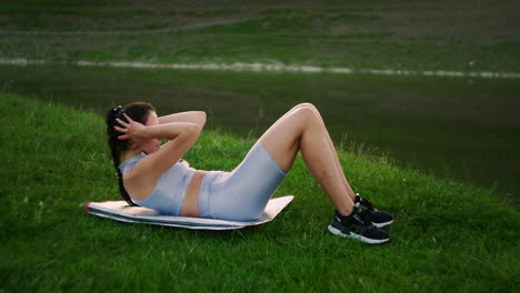 una mujer hace ejercicios para los músculos de la prensa acostada en la hierba en un parque cerca del lago. trabajar con los músculos abdominales. crear un cuerpo hermoso