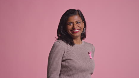 studio portrait of smiling mature woman proudly wearing pink clothing and breast cancer awareness ribbon against pink background