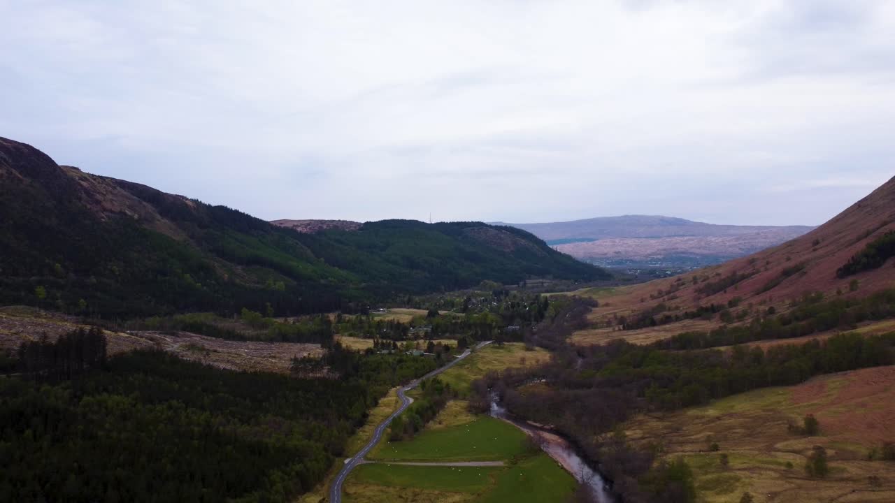 Baglæns droneoptagelse af vej og flod i de skotske Glens mod en skyet skotsk sommerhimmel