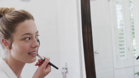 Happy-biracial-woman-applying-beauty-face-mask-looking-at-mirror-in-bathroom,-slow-motion