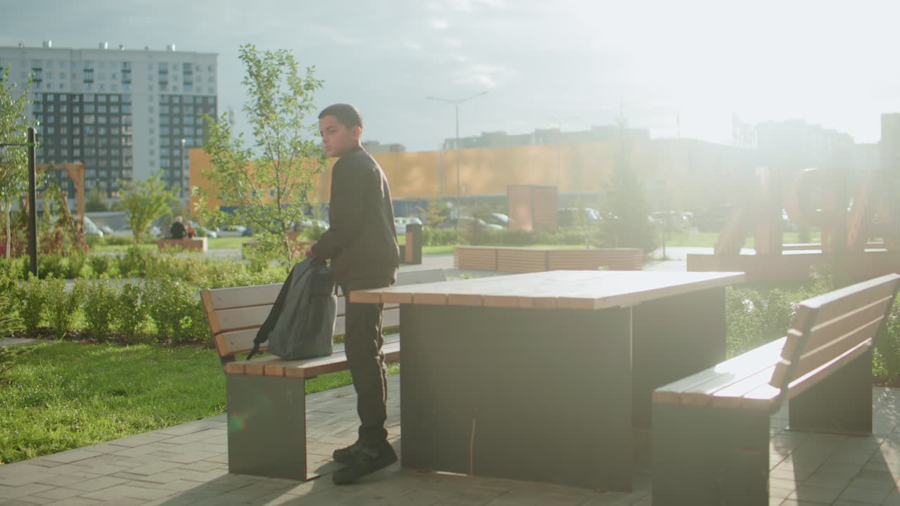 Student standing by bench outdoors zipping backpack after finishing study, preparing to walk away in bright daylight with green plants and modern buildings visible in background