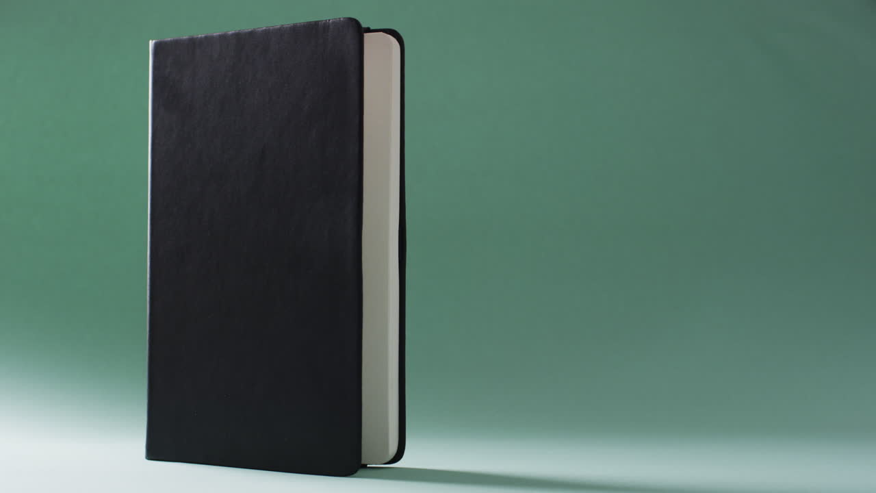 Close up of closed black book standing vertical with copy space on green background in slow motion
