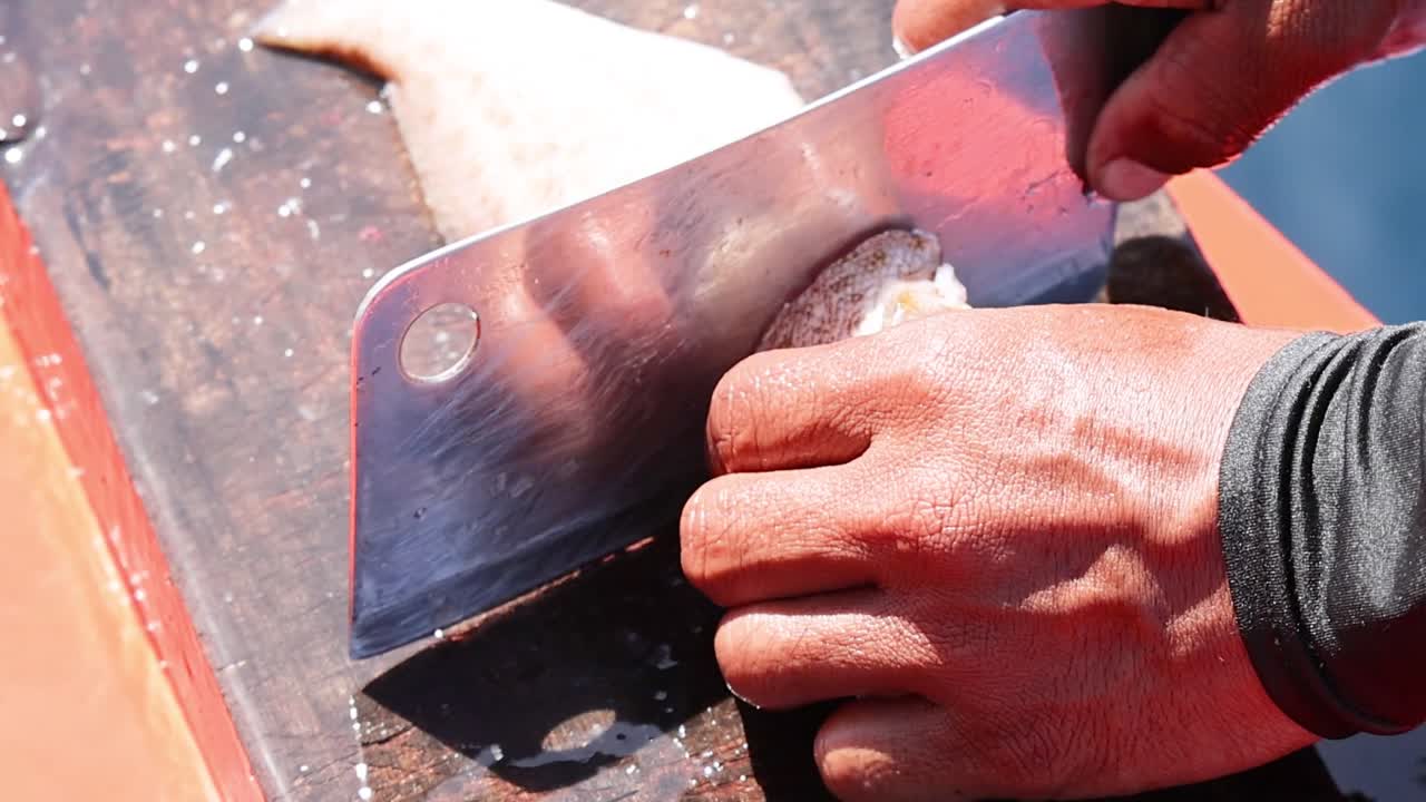 A close-up view of a person using a large metal cleaver to fillet fish
