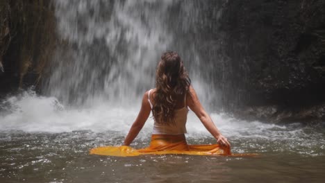 a slow motion shot of a young, attractive woman dancing in the water with a yellow skirt with a small waterfall behind her