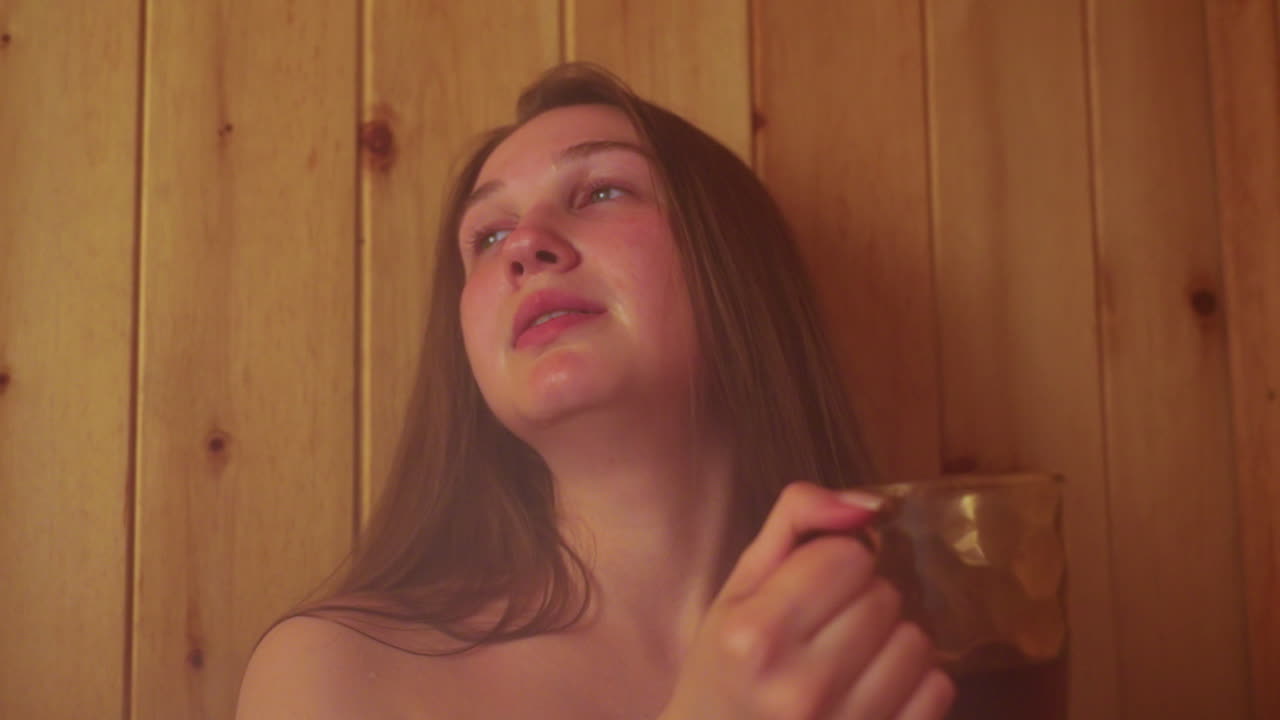 close up of woman sipping juice inside wooden sauna, leaning back against wall, eyes half closed in serene relaxation, wisps of steam rising around her as she enjoys warm herbal infusion