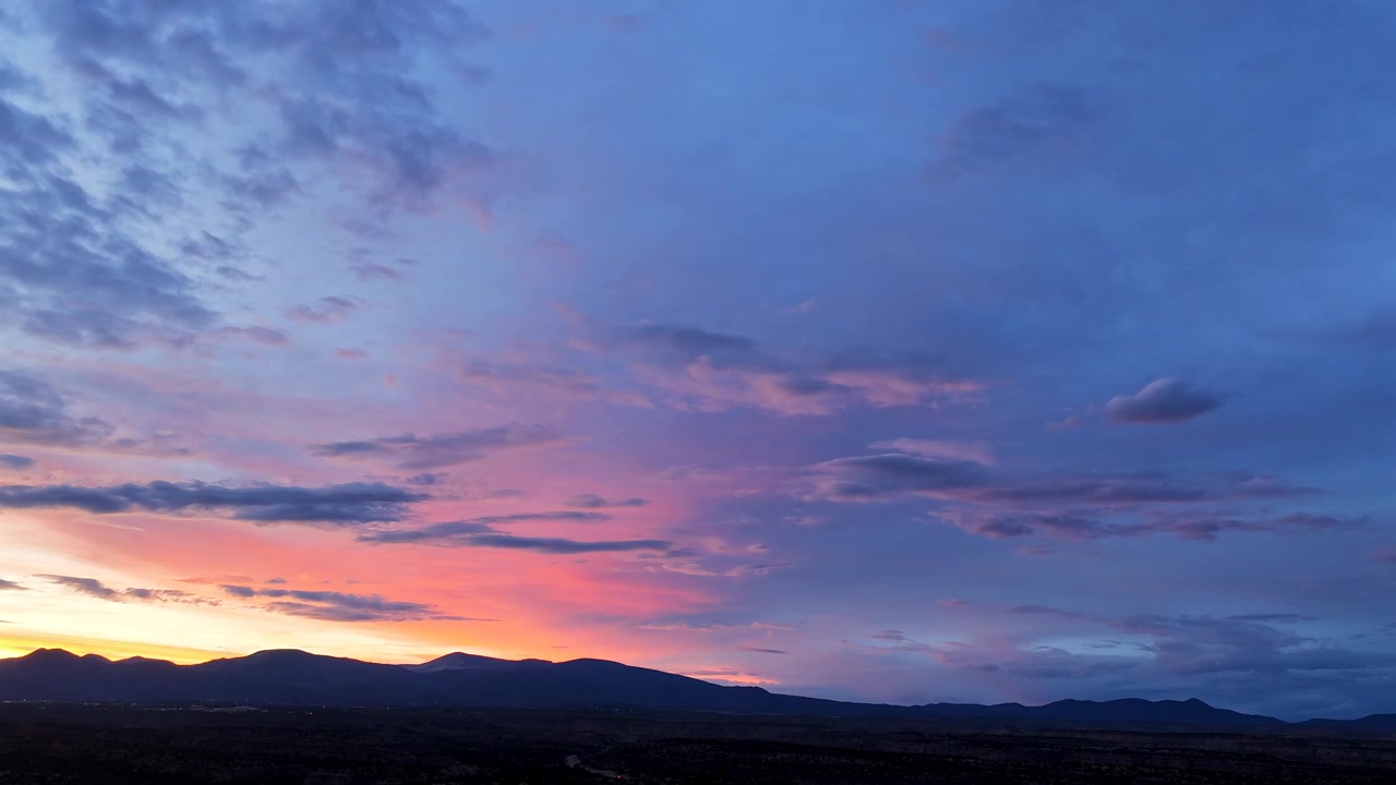 A drone timelapse capturing beautiful pastel sunset colors fading into twilight over a dark landscape. A distant road with moving car headlights is faintly visible along the horizon.