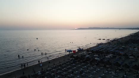 Puesta-De-Sol-Sobre-Una-Playa-Con-Sombrillas-Y-Gente-Caminando-Por-La-Orilla-Y-Sumergiéndose-En-El-Mar