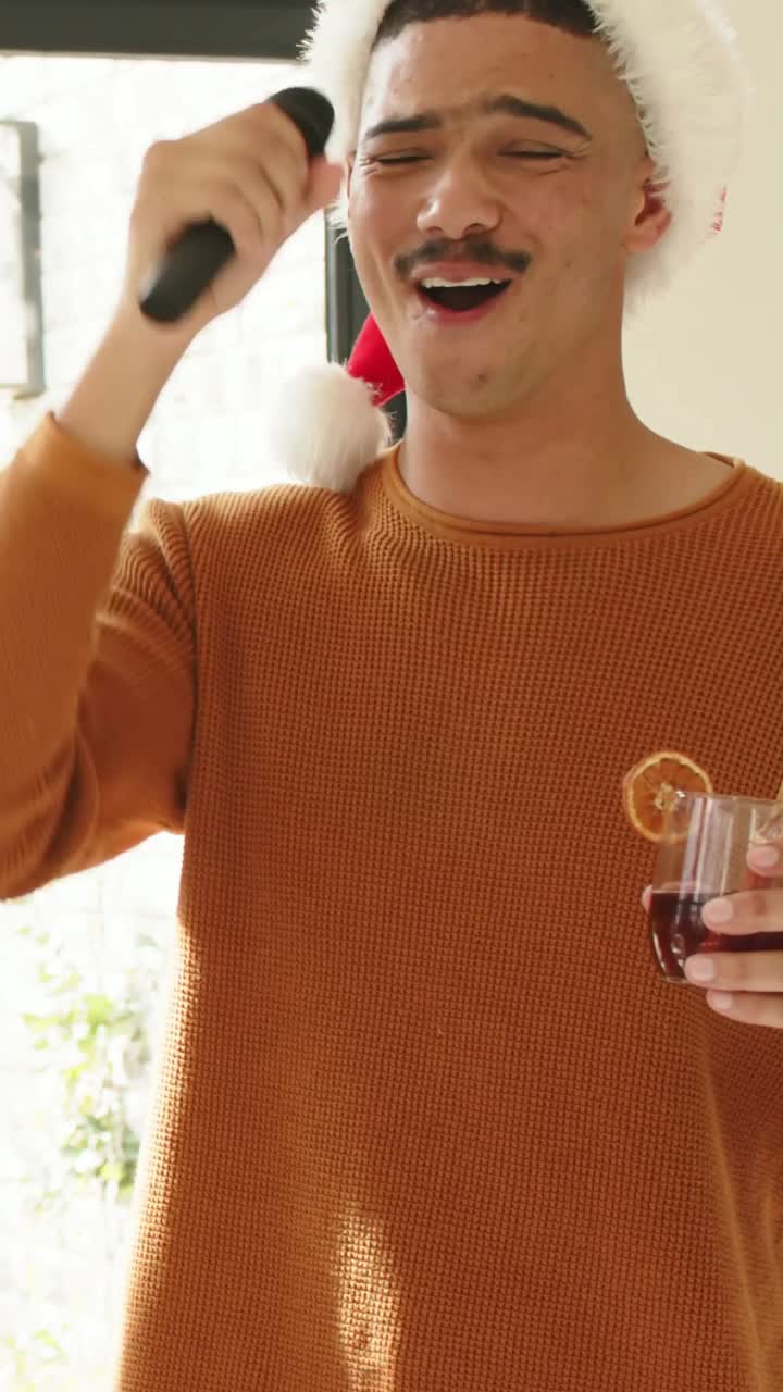 Vídeo vertical: Homem com gorro de Papai Noel e malha laranja, segurando microfone, canta perto da janela, brindando a convidado.