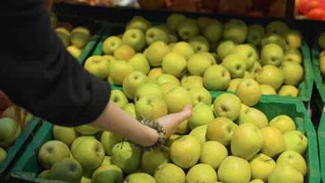 view of a hand wearing a black coat and bead bracelets picking up two apples from a crate in a grocery store