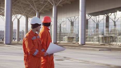 Two-construction-workers-in-orange-uniform-and-hardhats-examining-the-constructed-building-together.-Teamwork-concept