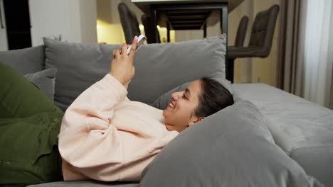 A-happy-brunette-girl-with-Brown-skin-in-a-light-sweater-and-green-pants-lies-on-a-gray-sofa-and-types-on-her-phone-happy-chatting-with-her-friends-online-at-home