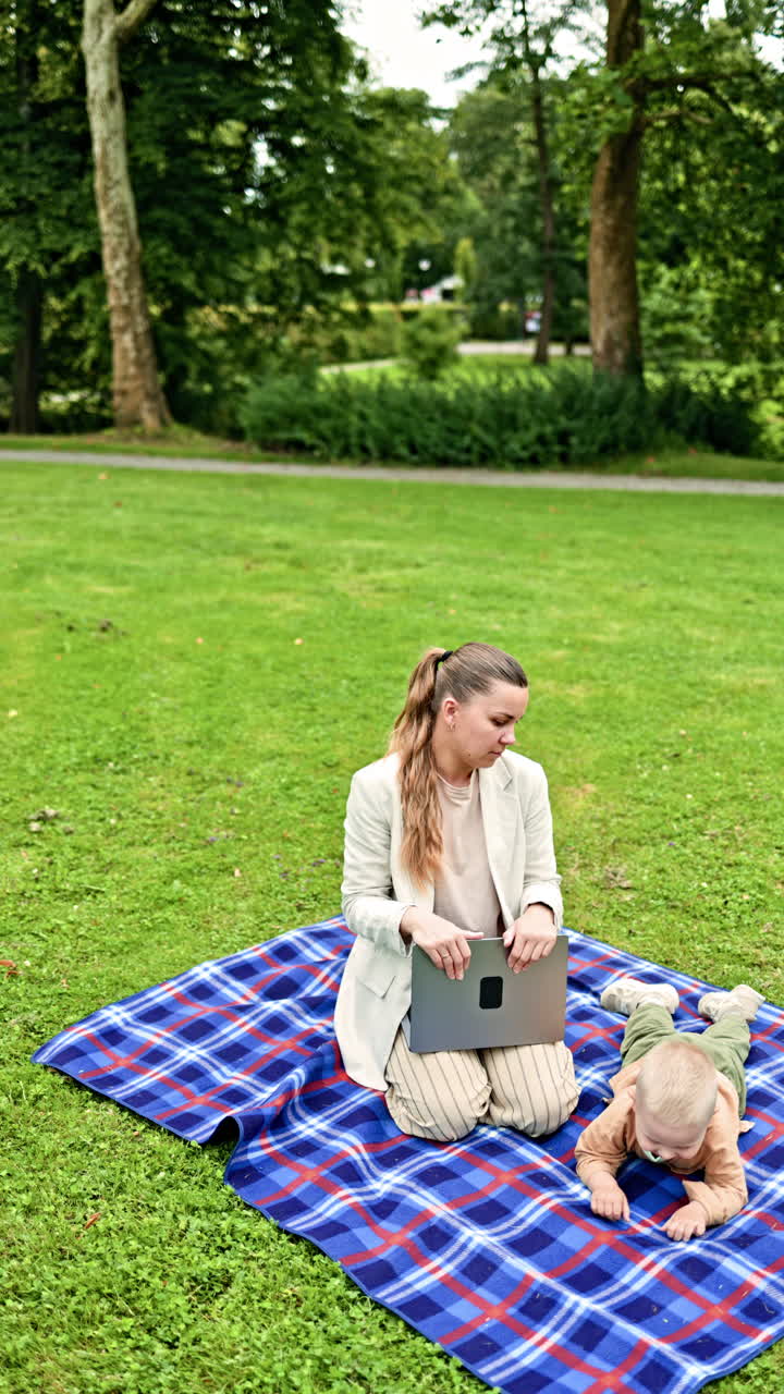 Koncentreret mor arbejder på computer siddende på græsset, mens lille søn løber i nærheden. Ung mor arbejder online på bærbar computer, mens hun går med søn i parken. Arbejdende mor på barselsorlov